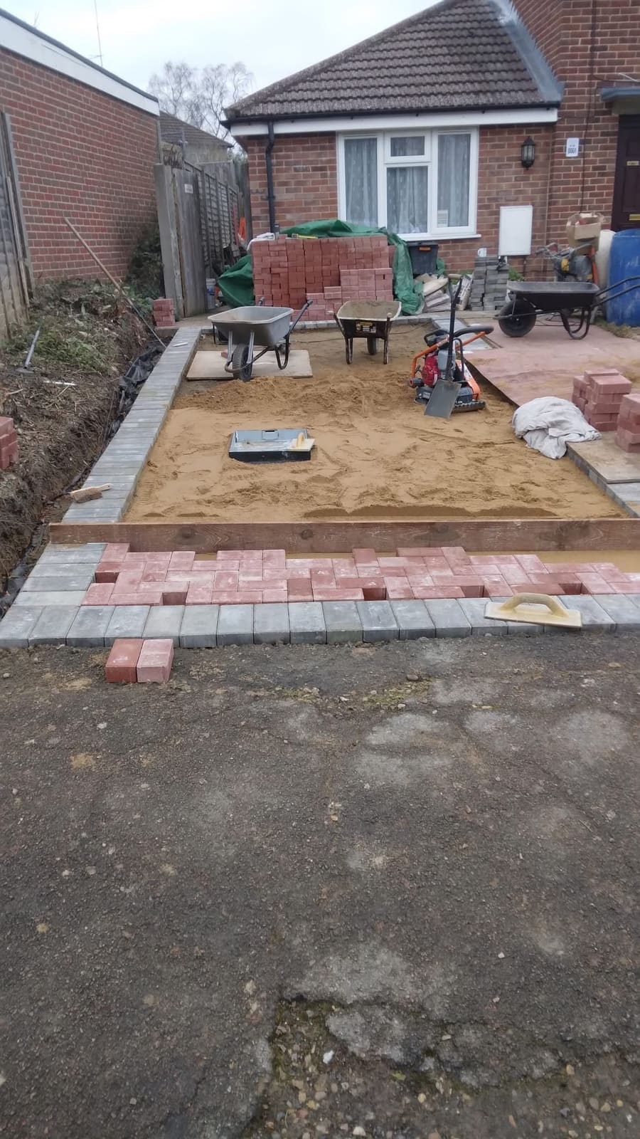 Block paving driveway under construction, showing the levelled sand screed, charcoal edging kerbs already laid and red block pavers stacked ready, with a plate compactor on site