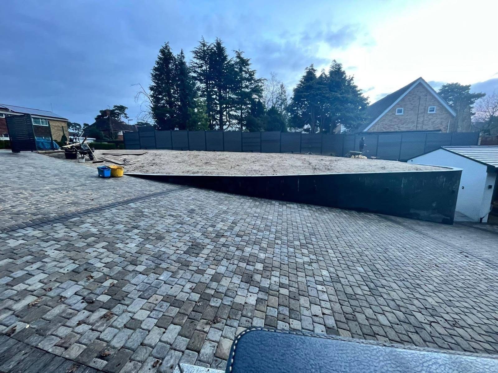 Large courtyard driveway laid in tumbled concrete setts, with a curved steel edge separating it from a sand-levelled area awaiting turf, and dark grey composite fence panels behind