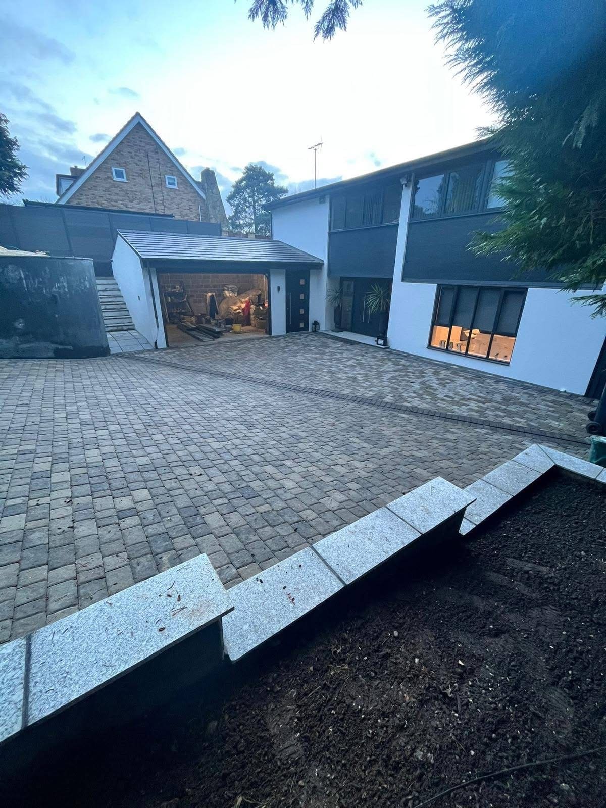 Tumbled sett driveway sweeping up to a rendered house and garage, edged with light granite kerbs against a newly dug planting bed