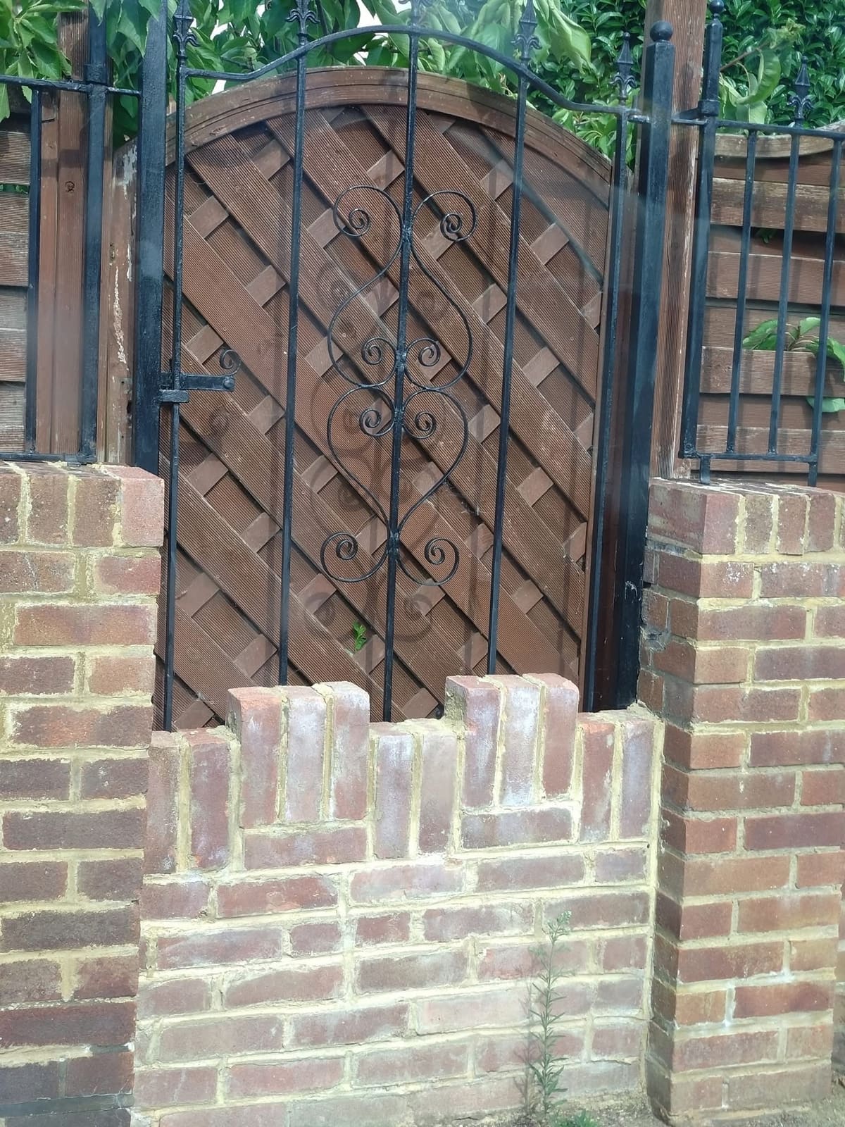 Wrought-iron gate with scrolled detail hung between red-brick piers, with a castellated brick wall and diagonal timber lattice panel behind