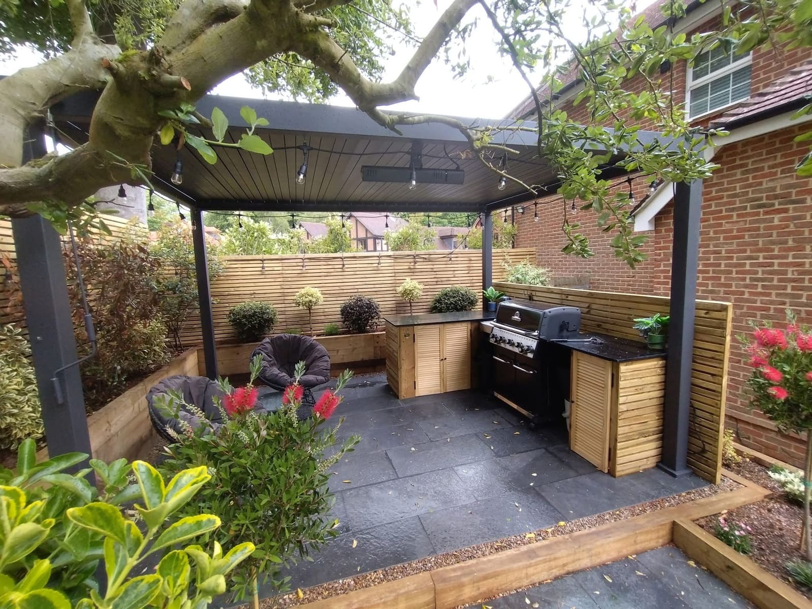Outdoor kitchen under an aluminium pergola, with a built-in gas barbecue, timber louvred cabinets, black granite worktop and slate paving, enclosed by slatted timber screens