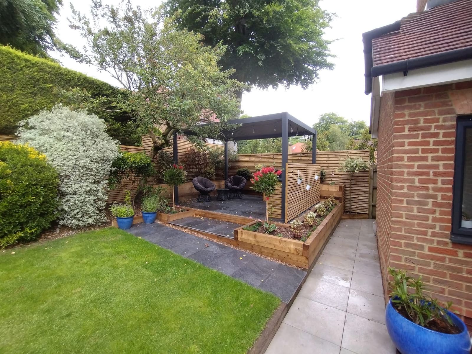 Rear garden with a striped lawn, slate paving path and a slatted timber screened seating area under an aluminium pergola, beside a red-brick house