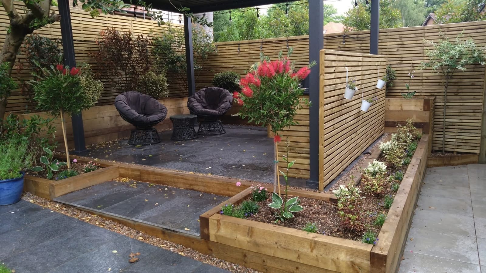 Garden seating area under a dark aluminium pergola with festoon lighting, framed by horizontal slatted timber screens and railway sleeper raised beds planted with bottlebrush and standard bay trees