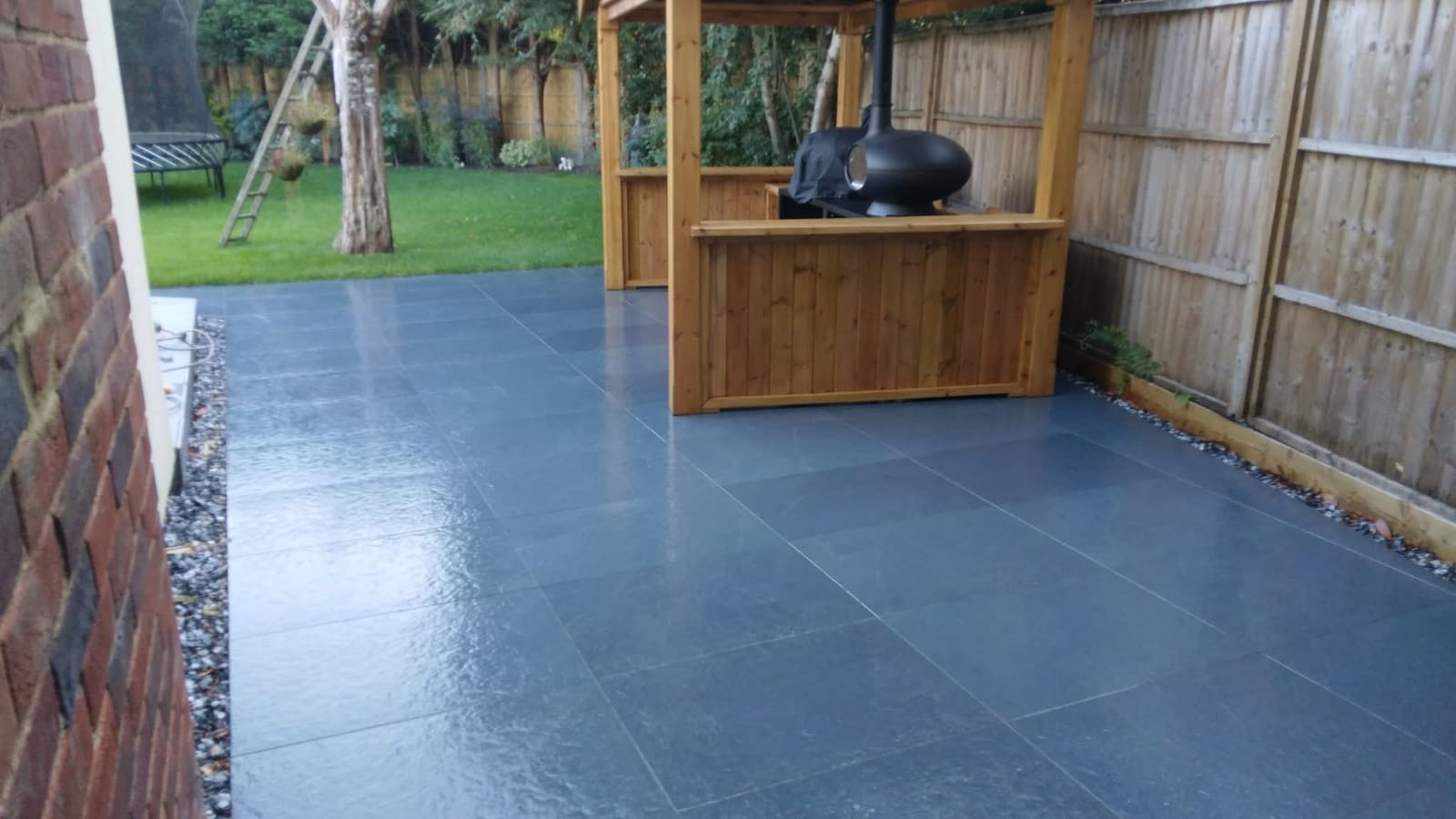 Charcoal large-format porcelain patio with a gravel drainage margin, leading to a timber garden bar with a pizza oven under a pitched shelter