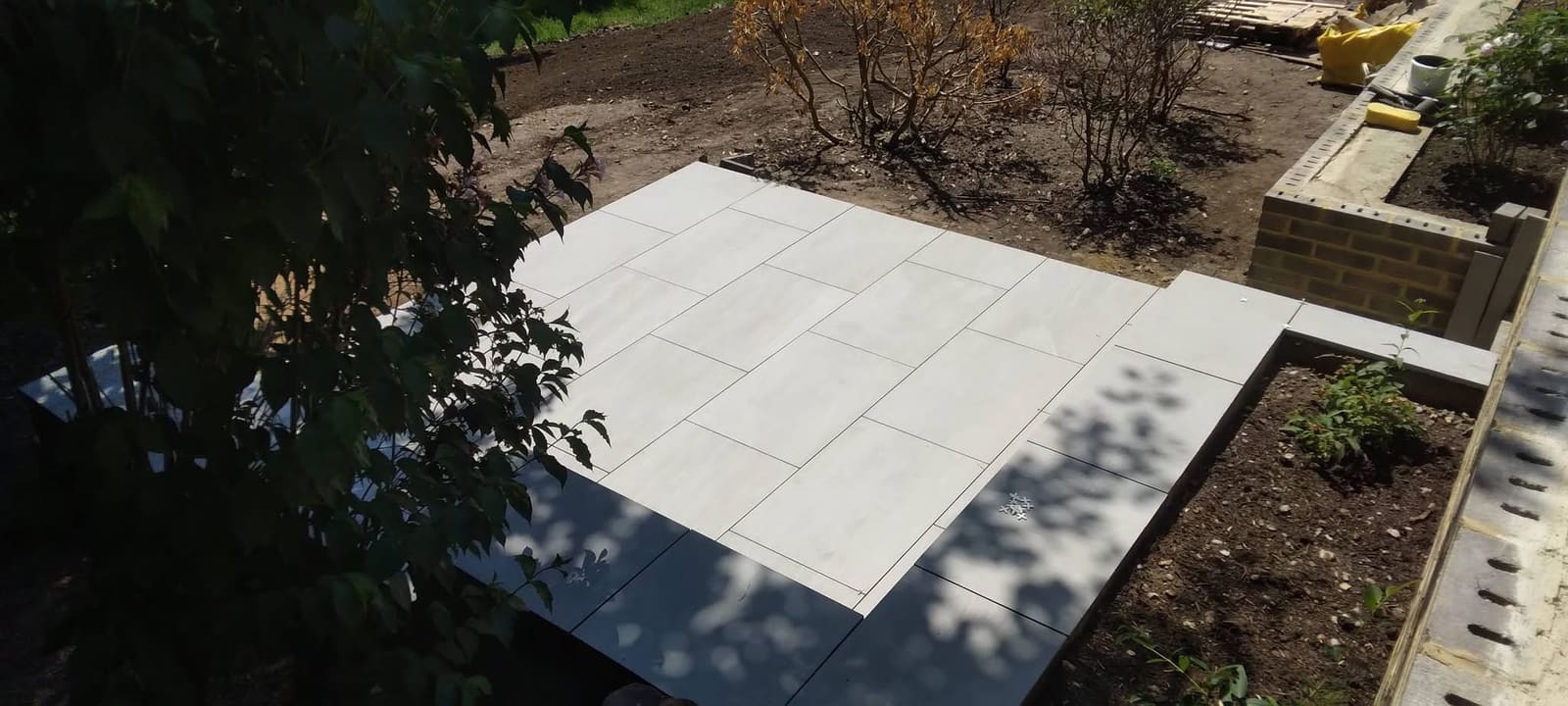 Light grey large-format porcelain paving part-laid with tile spacers still in place, beside a newly built yellow-brick raised planter