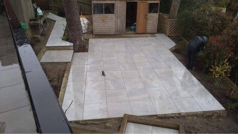 Light grey porcelain patio laid across a rear garden with timber sleeper edging and a step up to a garden shed