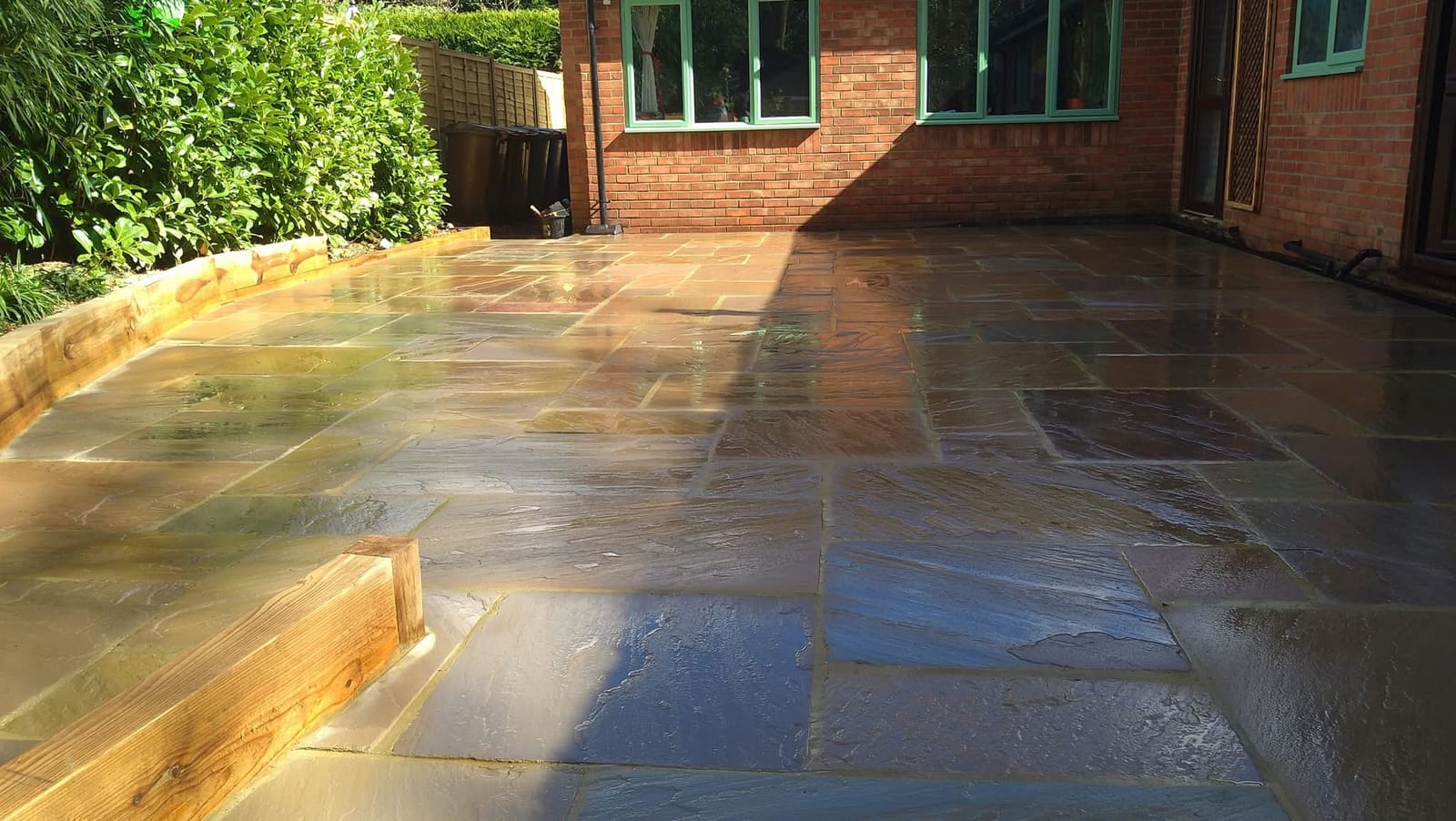 Indian sandstone patio in an autumn blend laid in a random rectangular pattern, wet after rain, with timber sleeper edging and a red-brick house with green window frames behind