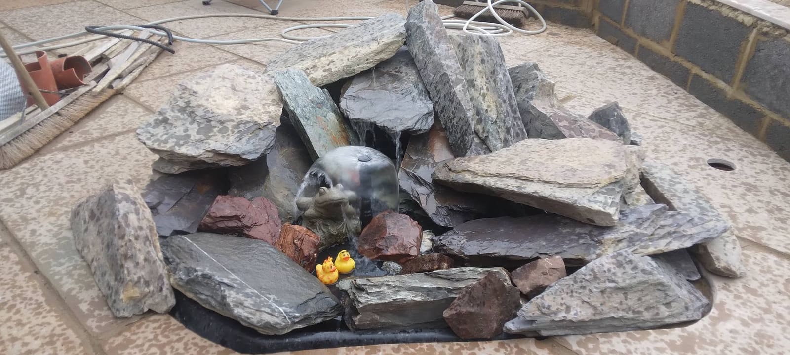 Stacked slate cascade water feature with a frog spout sitting in a reservoir tray on a patio, water running down the slate into the pool below
